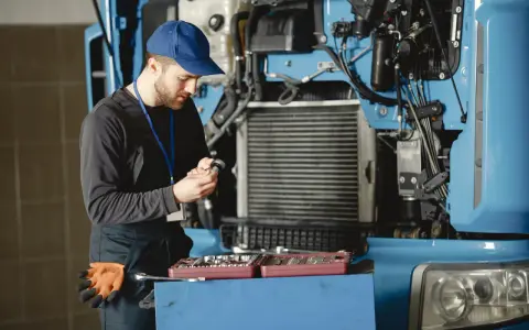 Mechanik w niebieskiej czapce i kombinezonie naprawia ciężarówkę, trzymając narzędzie.