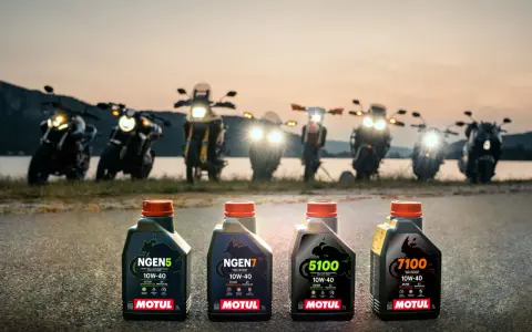 Cztery butelki oleju Motul do motocykla: NGEN5, NGEN7, 5100 i 7100, stoją na drodze o zachodzie słońca, za nimi widać rzędem motocykle.
