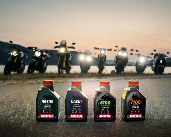 Cztery butelki oleju Motul do motocykla: NGEN5, NGEN7, 5100 i 7100, stoją na drodze o zachodzie słońca, za nimi widać rzędem motocykle.